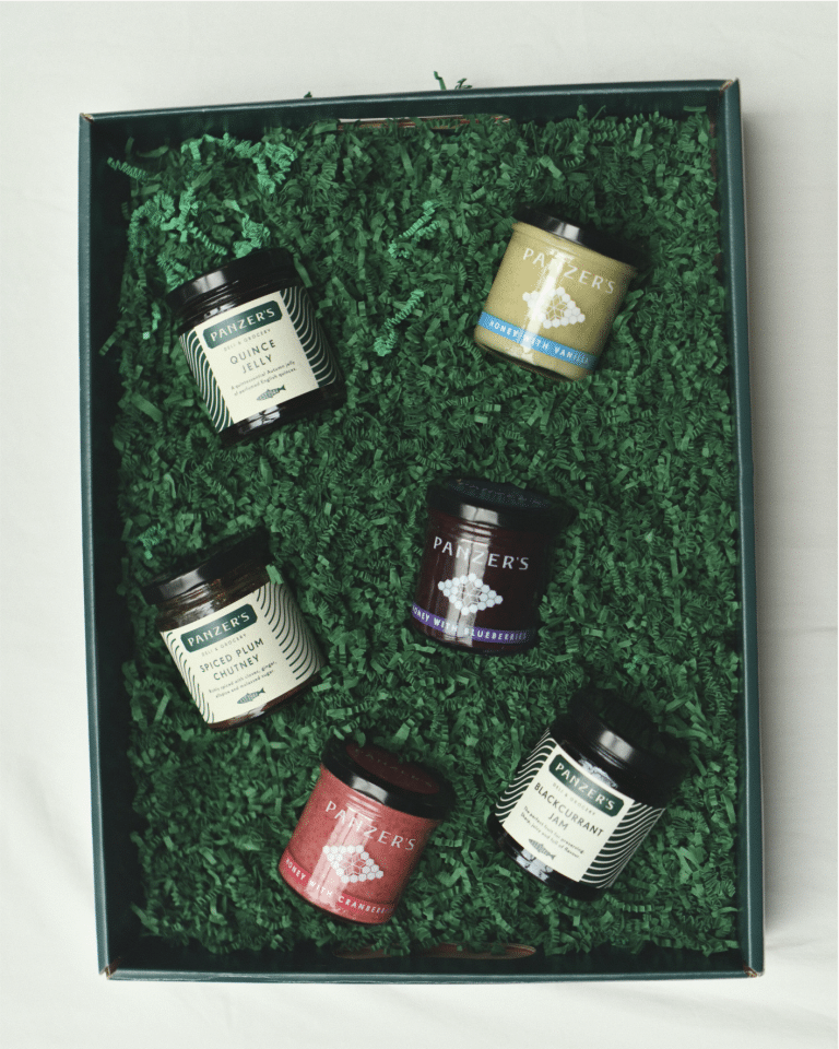 An assortment of Panzer’s preserves and honey, including jars of quince jelly, spiced plum chutney, honey with cranberries, and more, nestled in dark green shredded paper within an elegant gift box. Perfectly presented for gifting.
