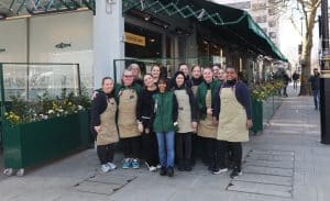 A group of women from the Panzer’s team stand together outside the shop, smiling at the camera. They are dressed in Panzer’s uniforms, including beige aprons and green jackets, with the store’s signage and flower arrangements visible in the background.