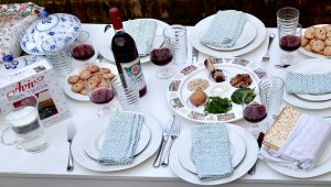 This image shows a beautifully set Passover Seder table. A Seder plate is at the center, featuring traditional symbolic foods such as a shank bone, an egg, bitter herbs, charoset, and parsley. Surrounding the Seder plate are plates with folded napkins, glasses filled with red wine, and a bottle of kosher wine. A box of Aviv Passover matzos is present, along with a covered stack of matzo on one of the plates. The table also includes plates of cookies, elegant blue-and-white covered serving dishes, and glassware. The setting is outdoors, with a white tablecloth creating a clean and festive presentation.