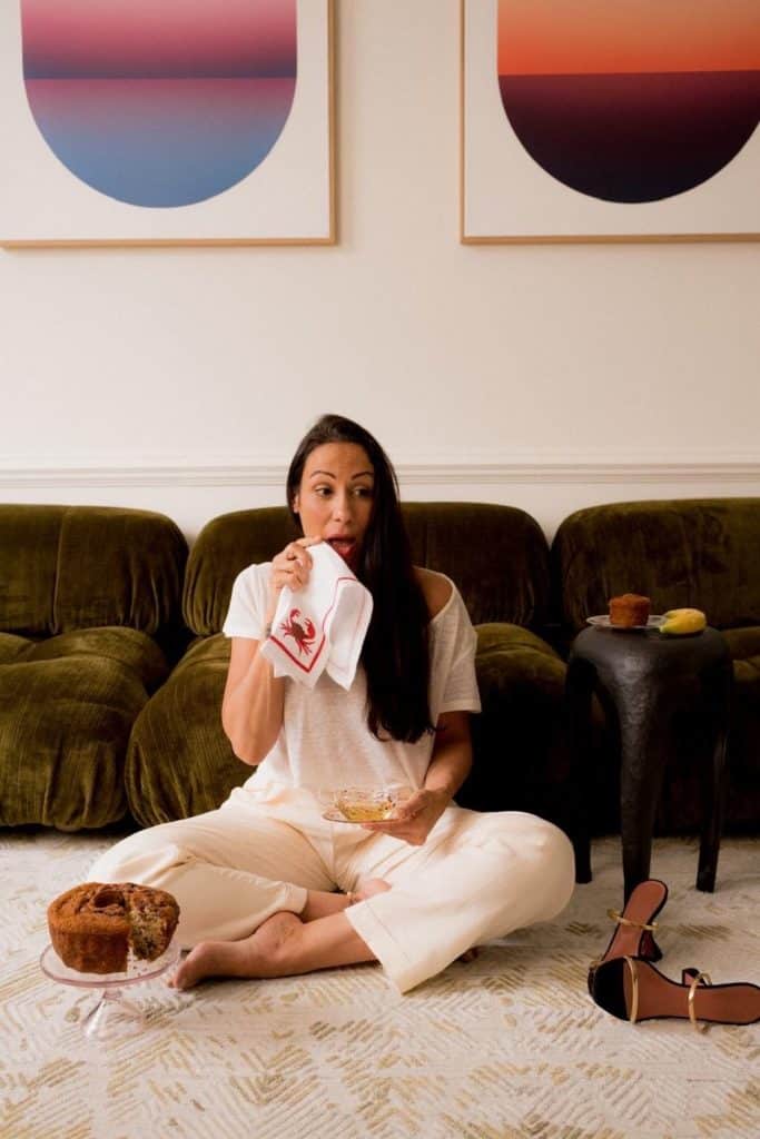 A woman with long dark hair, wearing a relaxed cream-colored outfit, sits barefoot on a patterned rug in a cozy living space. She holds a teacup and dabs her mouth with a white napkin featuring a red lobster design. A partially eaten cake sits on a glass stand beside her, while a black side table holds a muffin and a banana. Behind her, a plush olive-green sofa contrasts with two abstract sunset-themed artworks on the wall. A pair of black and gold high-heeled sandals are casually placed nearby.