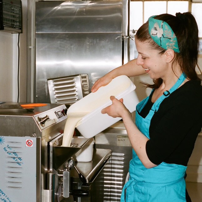 A woman wearing a turquoise apron and matching headband is pouring a creamy liquid into an ice cream or gelato machine in a professional kitchen. She is smiling as she works, surrounded by stainless steel equipment.