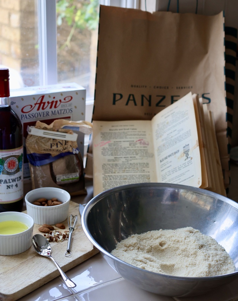 A cozy Passover baking scene set by a window, featuring a mixing bowl filled with almond flour. Surrounding it are ingredients and tools, including a small whisk, a spoon, chopped almonds, a dish of oil, and a bag of fine ground almonds. A vintage open cookbook, with handwritten notes, rests against a Panzer’s deli paper bag. Also present are a bottle of Palwin kosher wine and a box of Aviv Passover matzos, suggesting a traditional holiday recipe in the making.
