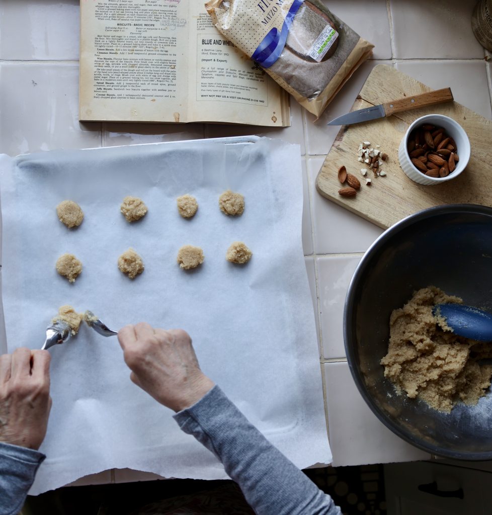 An overhead view of a Passover baking session, where hands carefully shape small almond-based biscuits onto parchment paper. The scene includes a mixing bowl filled with dough, a wooden board with chopped almonds and a small knife, and a vintage cookbook open to a classic recipe. A bag of fine ground almonds rests nearby, reinforcing the traditional nature of the bake. The setting is warm and nostalgic, evoking family traditions passed down through generations.