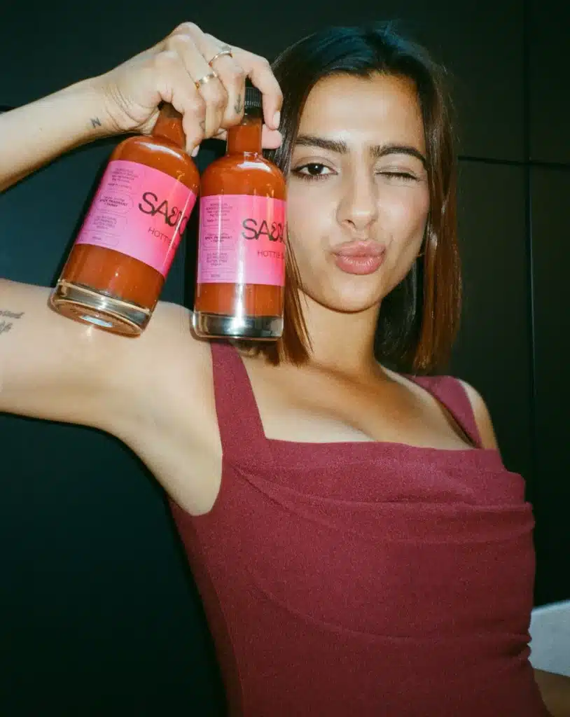 Woman in red dress holds two glass bottles of saucey dressing with red liquid inside while winking at the camera
