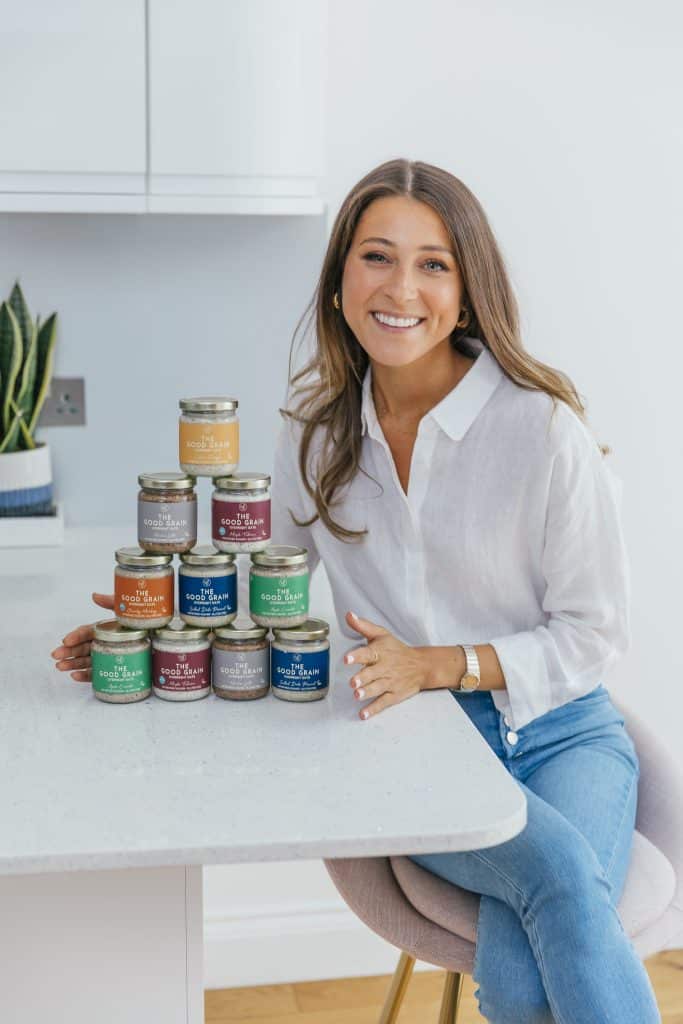 A woman with long brown hair, wearing a white blouse and blue jeans, is sitting at a white kitchen counter, smiling at the camera. In front of her is a neatly stacked pyramid of glass jars labeled "The Good Grain" in various colors, suggesting different flavors or varieties. The setting is bright and modern, with a plant visible in the background.