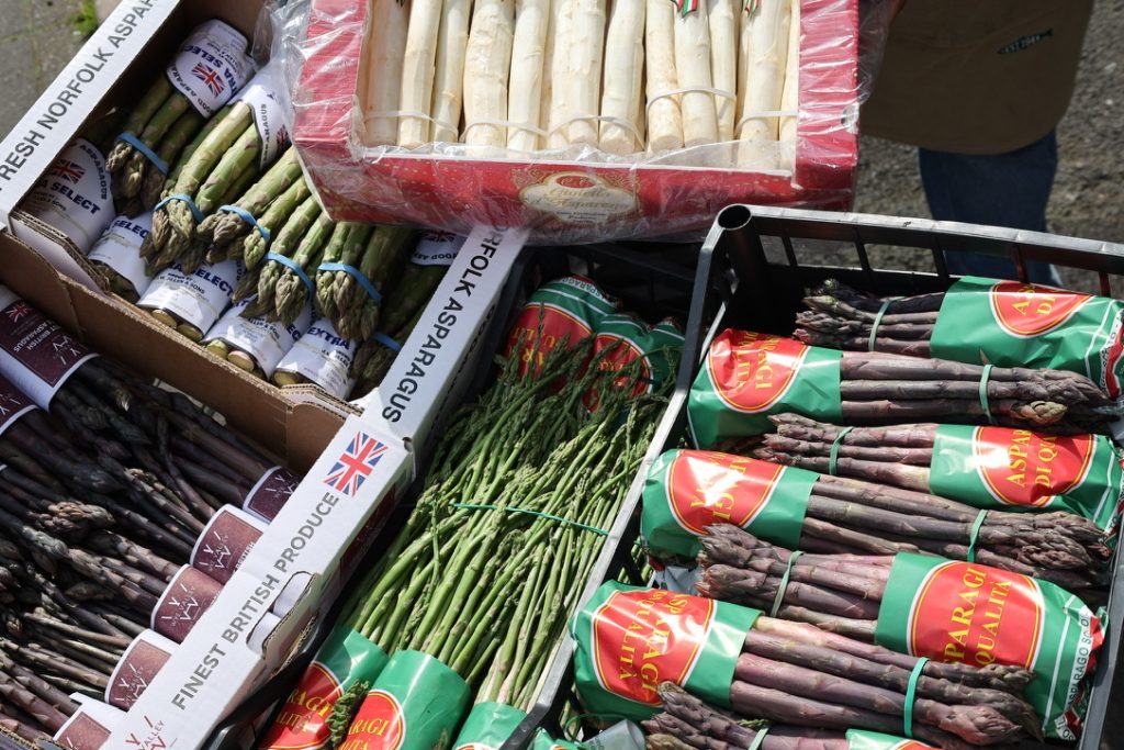 A colourful outdoor market display of various types of asparagus, including green, white, wild, and purple varieties. The asparagus is neatly bundled and packed in boxes and crates, some labelled as British produce and others with Italian branding.