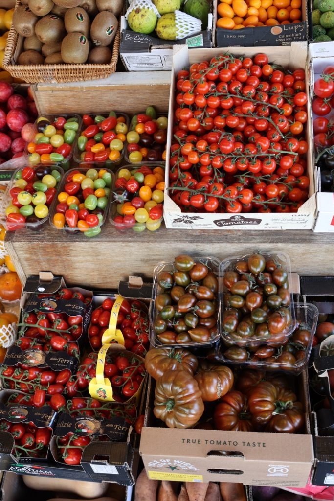 Assorted colourful tomatoes in crates, including yellow cherry, orange plum, red beefsteak, and dark green-black varieties, all neatly arranged at a market stall.