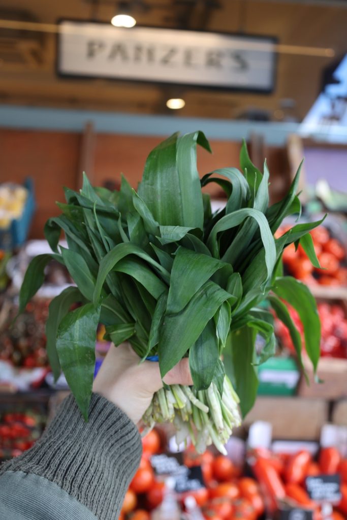 Hand holding a bunch of wild garlic in front of panzer's fruit & veg display outside