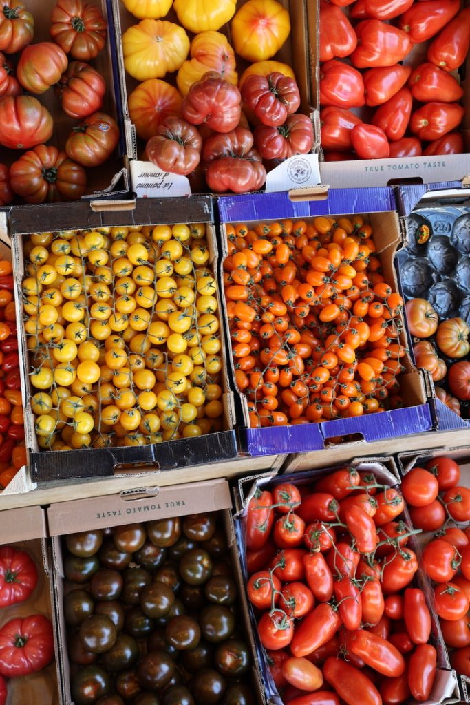 Assorted colourful tomatoes in crates, including yellow cherry, orange plum, red beefsteak, and dark green-black varieties, all neatly arranged at a market stall.